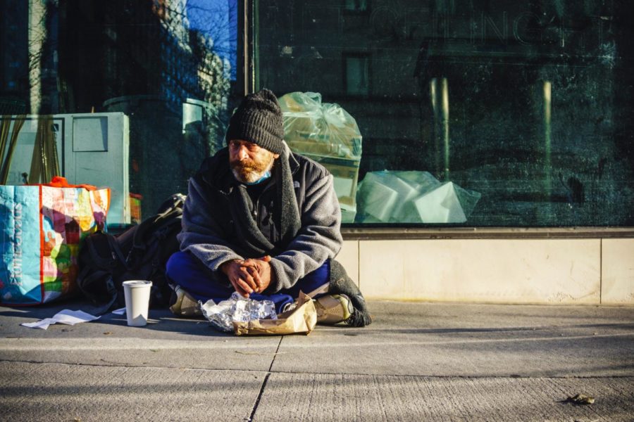 A homeless man sits on the ground on a New York City sidewalk, enjoying a coffee and a small bite to eat. This is an example of how public spaces are currently being used by the homeless community. 