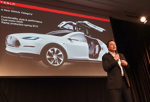 Elon Musk addresses his audience at a Tesla convention, his net worth rising every minute. 