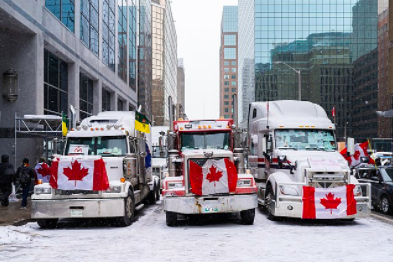 This is a photo of trucks with the Canadian flag in front blocking the roads as a sign of protest against the mandate in the streets of Ottawa. There are also many cars within this line amongst the trucks which shows how united many people are regarding this issue.