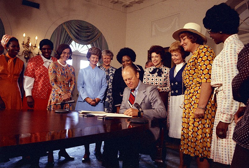 Representative Bella Abzug (D-NY) proudly looks over President Gerald R. Ford’s shoulder as he signs the Proclamation on Women’s Equality Day, along with fellow Representatives Yvonne Brathwait Burke (D-CA), Barbara Jordan (D-TX), Elizabeth Holtzman (D-NY), Marjorie S. Holt (R-MD), Leonor K. Sullivan (D-MO), Cardiss Collins (D-IL), Corinne C. Boggs (D-LA), Margaret M. Heckler (R-MA), and Shirley Chisholm (D-NY).   