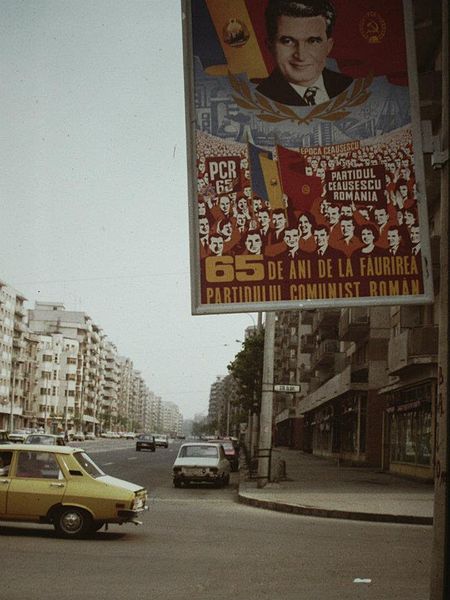 Pictured is a propaganda poster on the streets of Bucharest. The poster declares “65 years since the creation of the Romanian Communist Party!” There is a crowd depicted behind the text, with people holding banners that state “Ceausescu’s Epoch” and “PCR”, which stands for Romanian Communist Party. At the top is Romania’s communist-era flag, displaying Ceausescu’s face, the coat of arms and the flag. (Photo Credit: Scott Edelman, Public domain, via Wikimedia Commons)