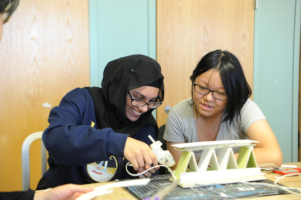 Students construct a bridge out of cardboard and hot glue, preparing to compete with the rest of the class to see which bridge supports the most weight. 