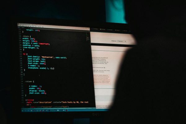 A shadowy figure is writing style code for a website, which closely resembles hacking, because there is colorful text on a screen. (Photo Credit: Mika Baumeister / Unsplash)