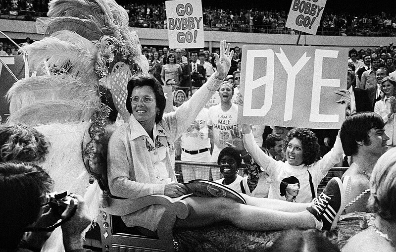 Before the start of the Battle of the Sexes against Riggs, Billie Jean King is pictured here on a litter and carried by four men to the tennis court. (Photo Credit: Associated Press, Public domain, via Wikimedia Commons)