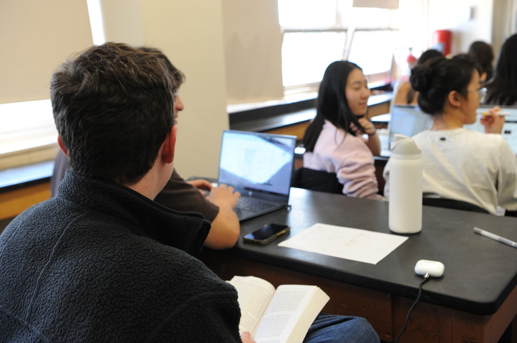 Here is an over the shoulder photograph of Bronx Science students in Advanced Placement Environmental Science working on their laptops, with a student reading a book with his airpods and his classmate's phone beside him.